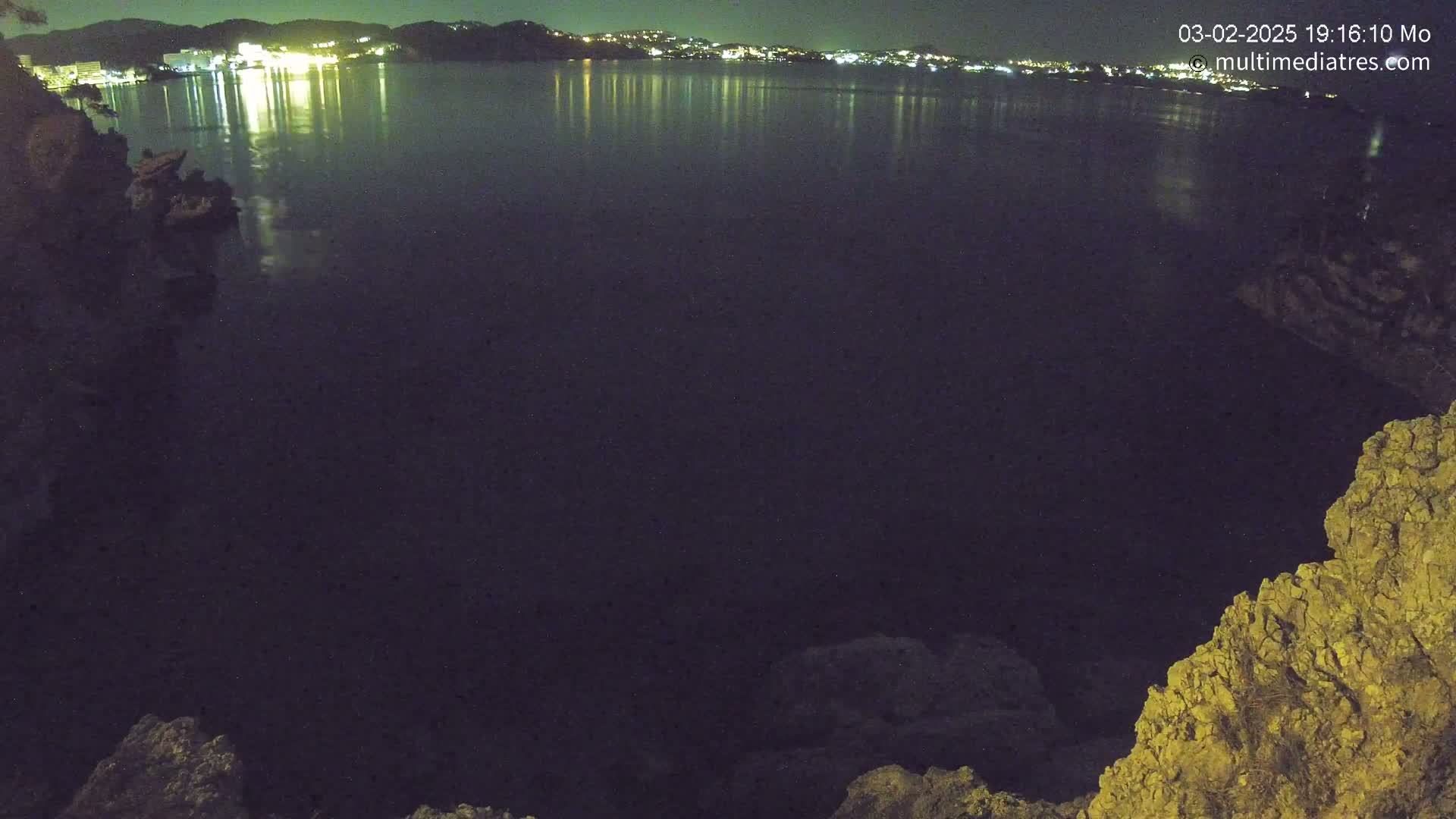 A nighttime view of a dark body of water reflecting the lights of a town across the water, seen from a rocky vantage point.