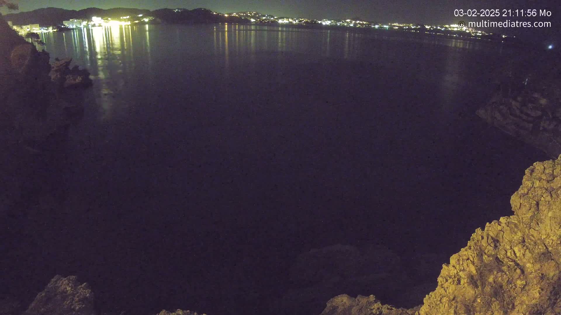 A nighttime view shows a dark body of water reflecting the lights of a distant town, with rocky terrain visible in the foreground.
