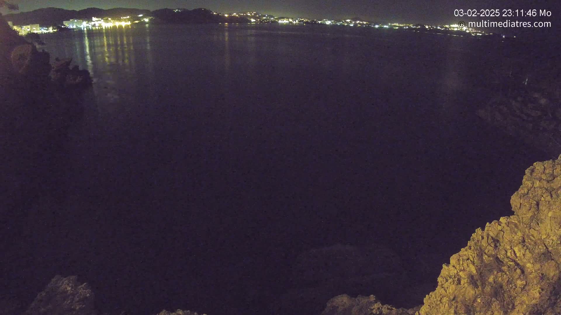 A nighttime view of a dark body of water reflecting the lights of a distant town, seen from a rocky vantage point.