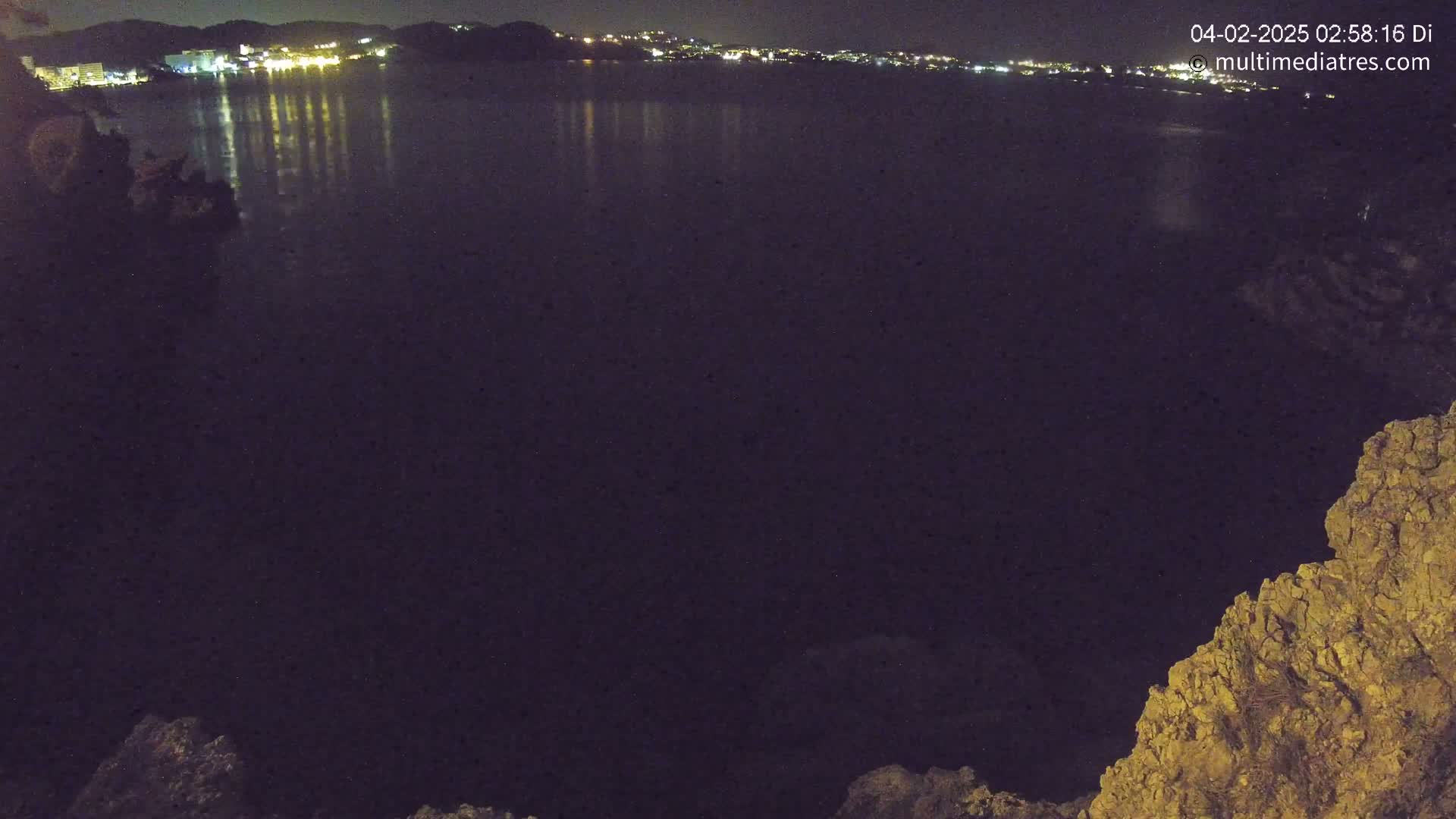 A nighttime view of a calm body of water reflecting the lights of a town on the opposite shore, seen from a rocky vantage point.