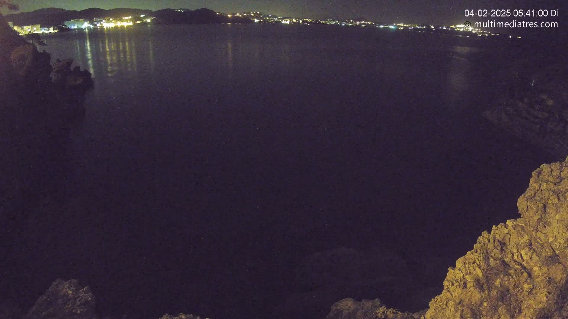 A nighttime view of a dark body of water reflecting the lights of a distant town, seen from a rocky overlook.