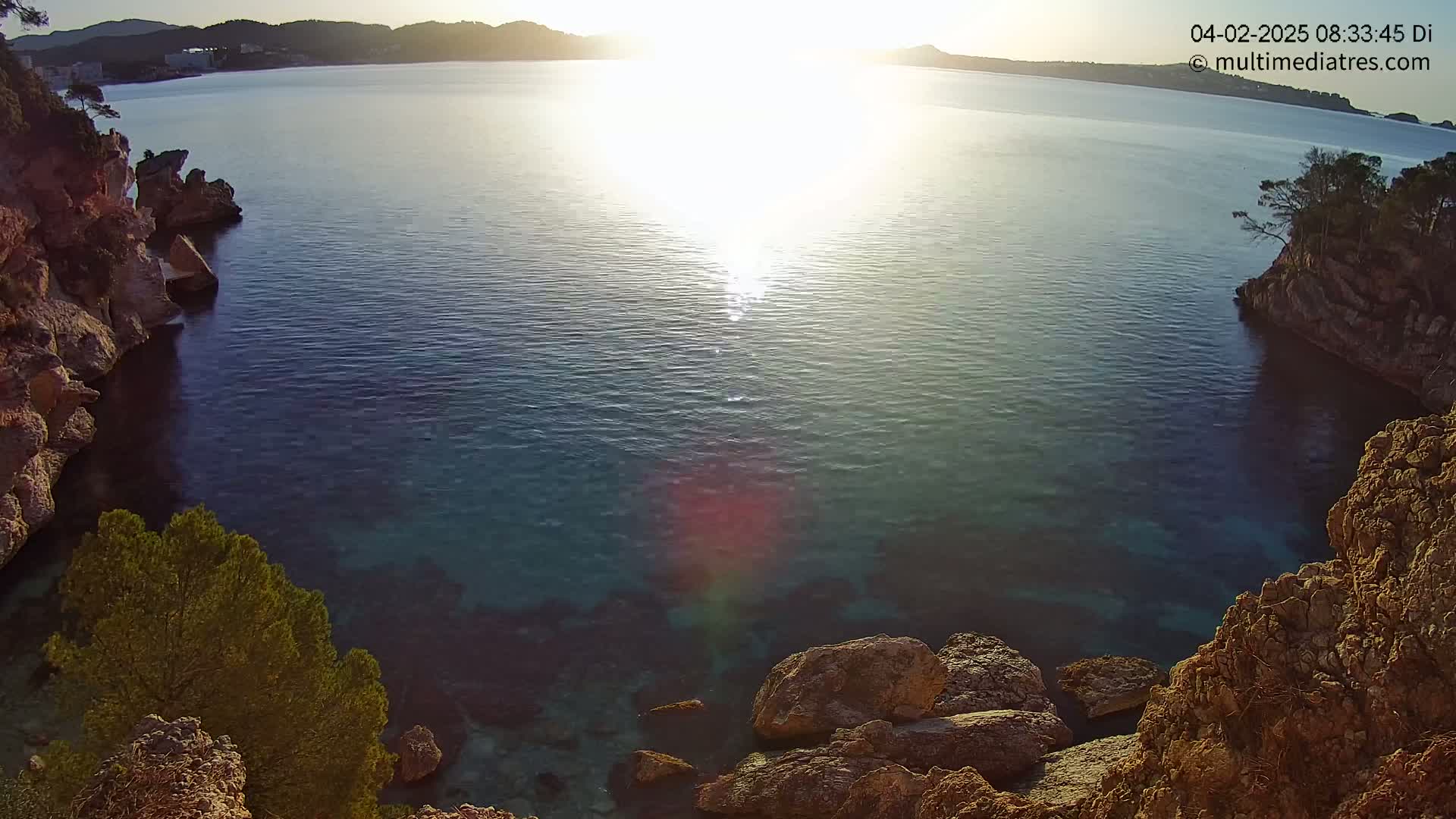 Rocky cliffs frame a calm, sunlit cove of clear blue water, with a distant shoreline visible under a bright, possibly sunrise or sunset sky.