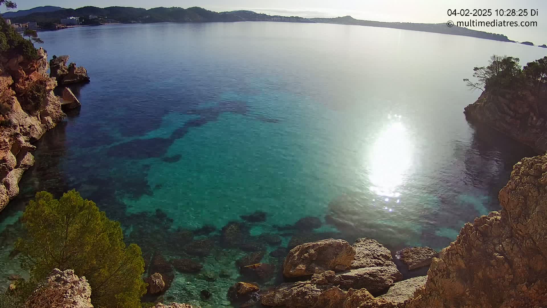 Clear, bright sunlight shines on a calm, teal-colored ocean cove surrounded by rocky cliffs and vegetation.