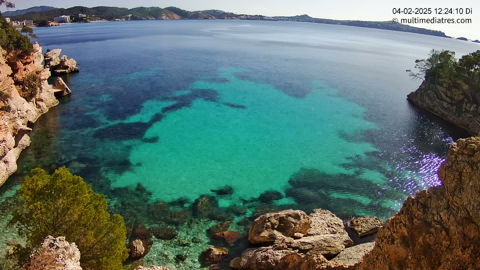 A cove of clear, teal water is surrounded by rocky, tan-colored cliffs and vegetation under a sunny sky.