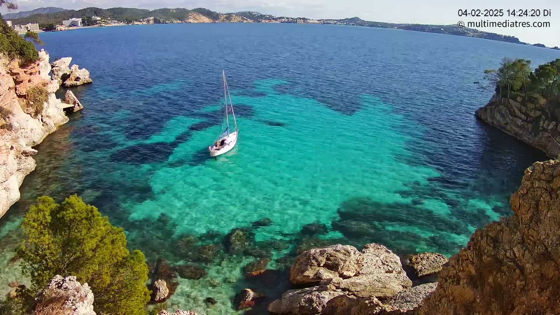 A sailboat sits in a calm, clear turquoise cove surrounded by rocky cliffs on a sunny day.