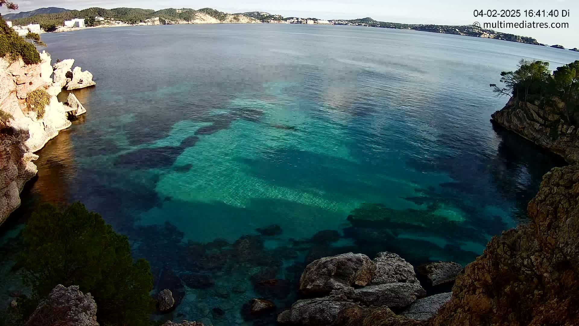 Clear, teal water fills a cove between rocky cliffs on a sunny day, with a distant coastline visible.
