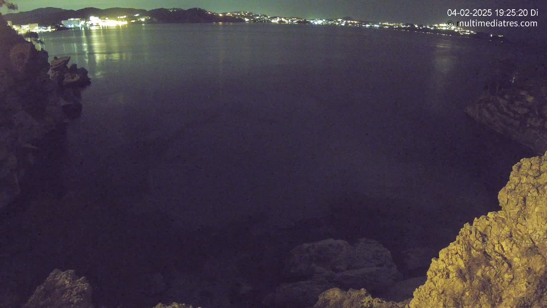 A nighttime view of dark water reflecting city lights across a bay, seen from a rocky outcrop.