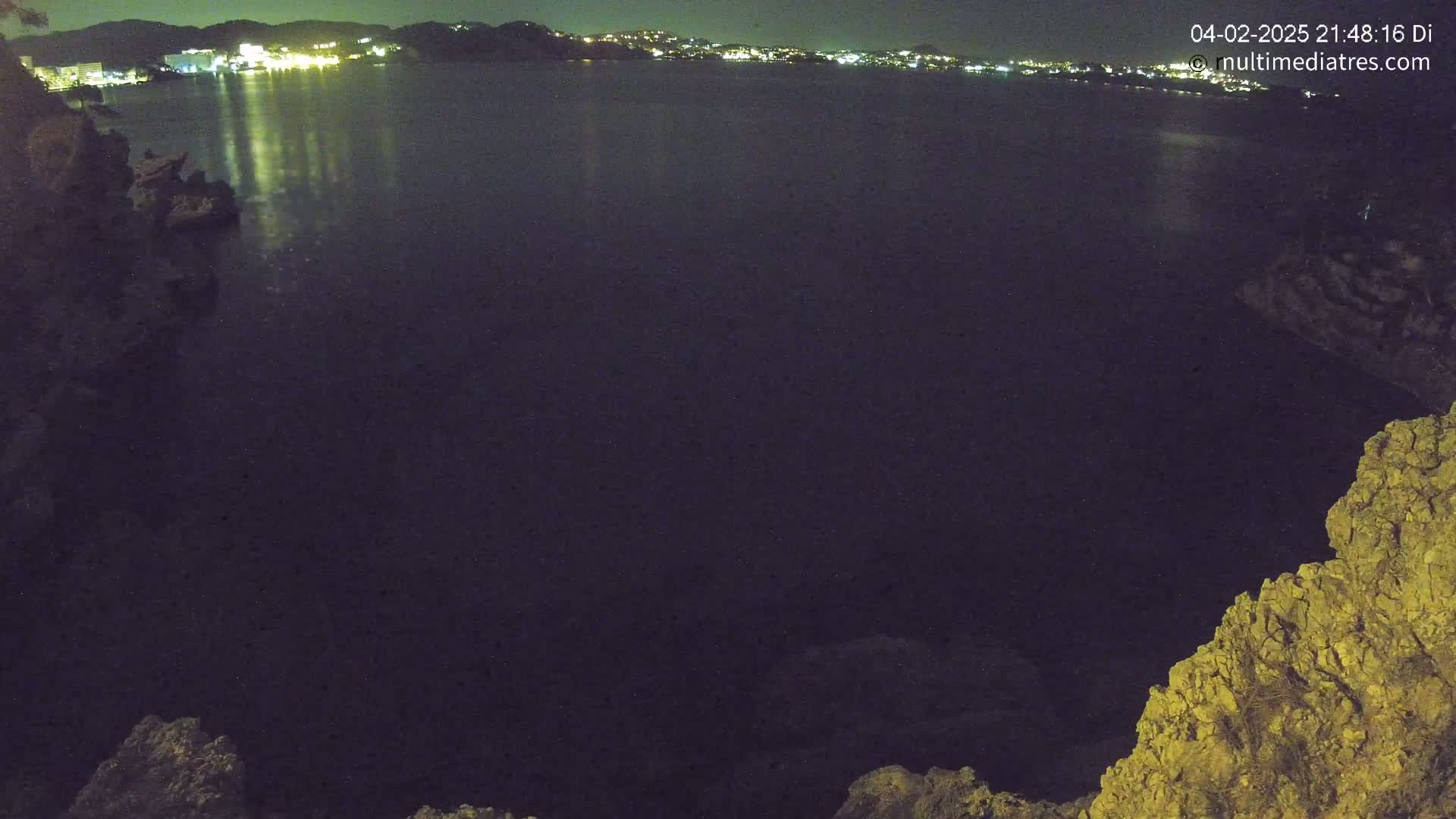 A nighttime view of a dark body of water reflecting the lights of a town on the opposite shore, seen from a rocky cliff.