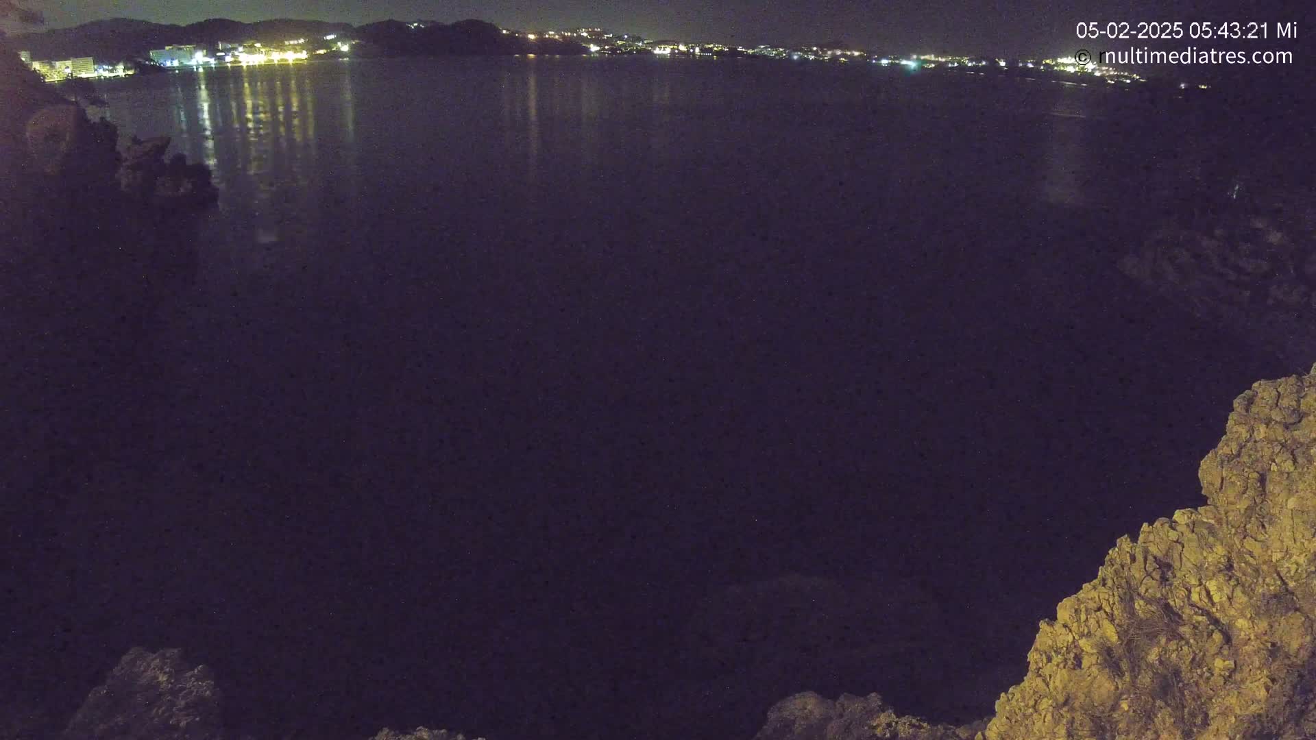 A nighttime view of a dark body of water reflecting the lights of a distant town, seen from a rocky vantage point.