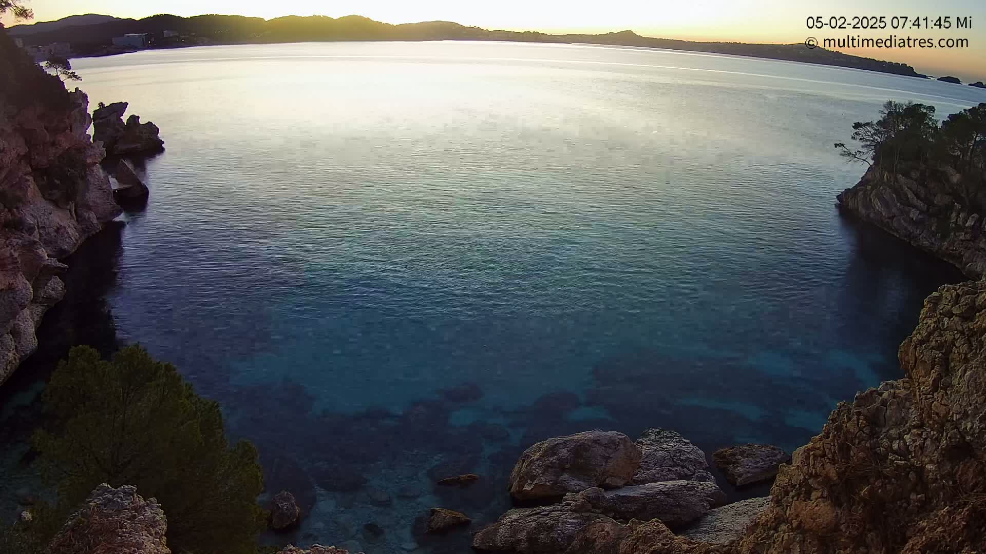 Calm, clear, blue water fills a cove surrounded by rocky cliffs and sparse vegetation under a sunrise sky.