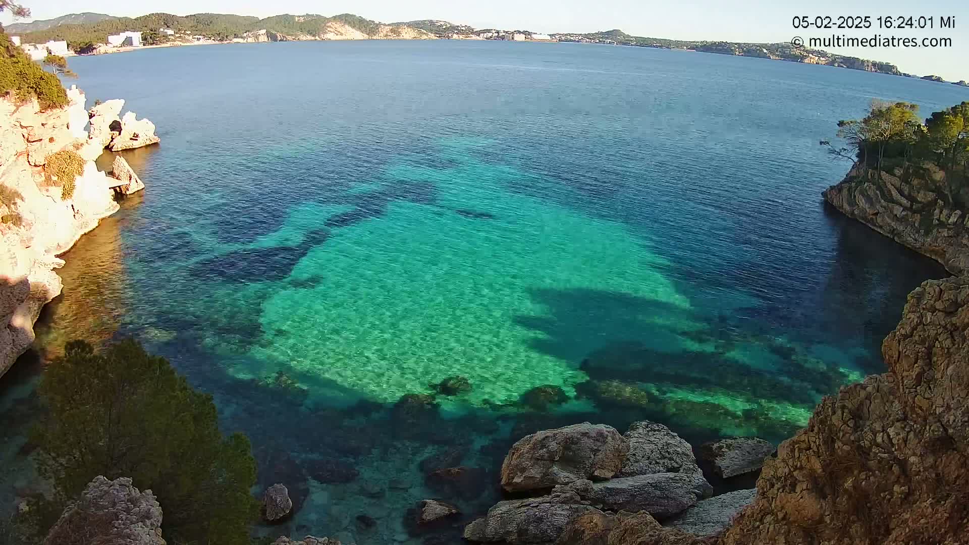 Clear, turquoise water fills a cove between light-colored rocky cliffs on a sunny day, with a distant coastline visible in the background.