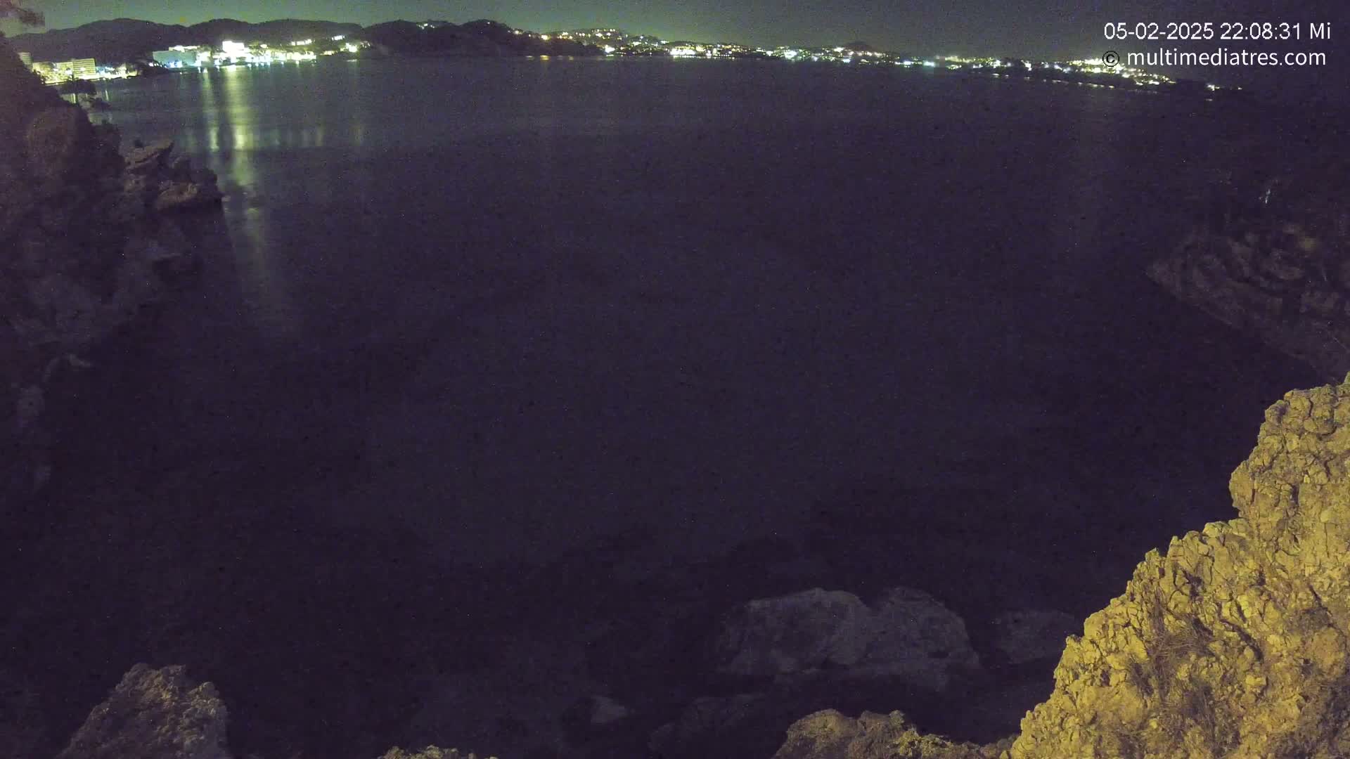 A nighttime view of a dark body of water reflecting the lights of a distant town, seen from a rocky, elevated vantage point.