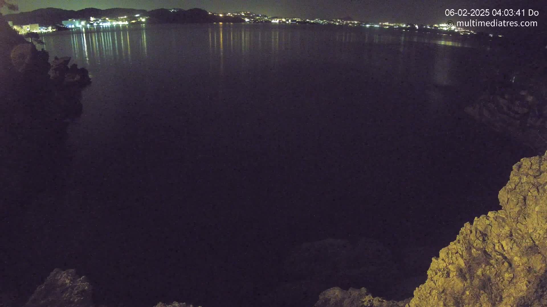 A nighttime view of a dark body of water reflecting the lights of a city in the distance, with rocky terrain visible in the foreground under a clear sky.