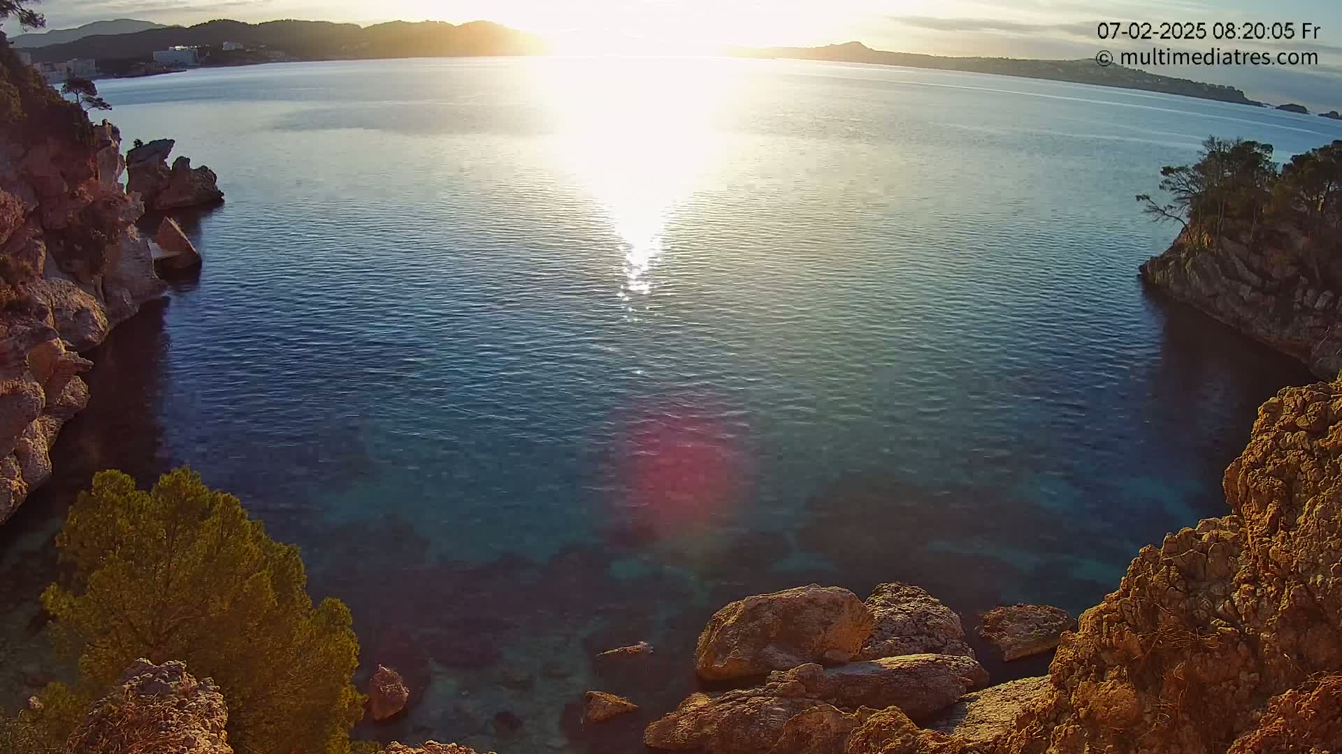 Rocky cliffs enclose a calm, clear cove of blue water at sunset.