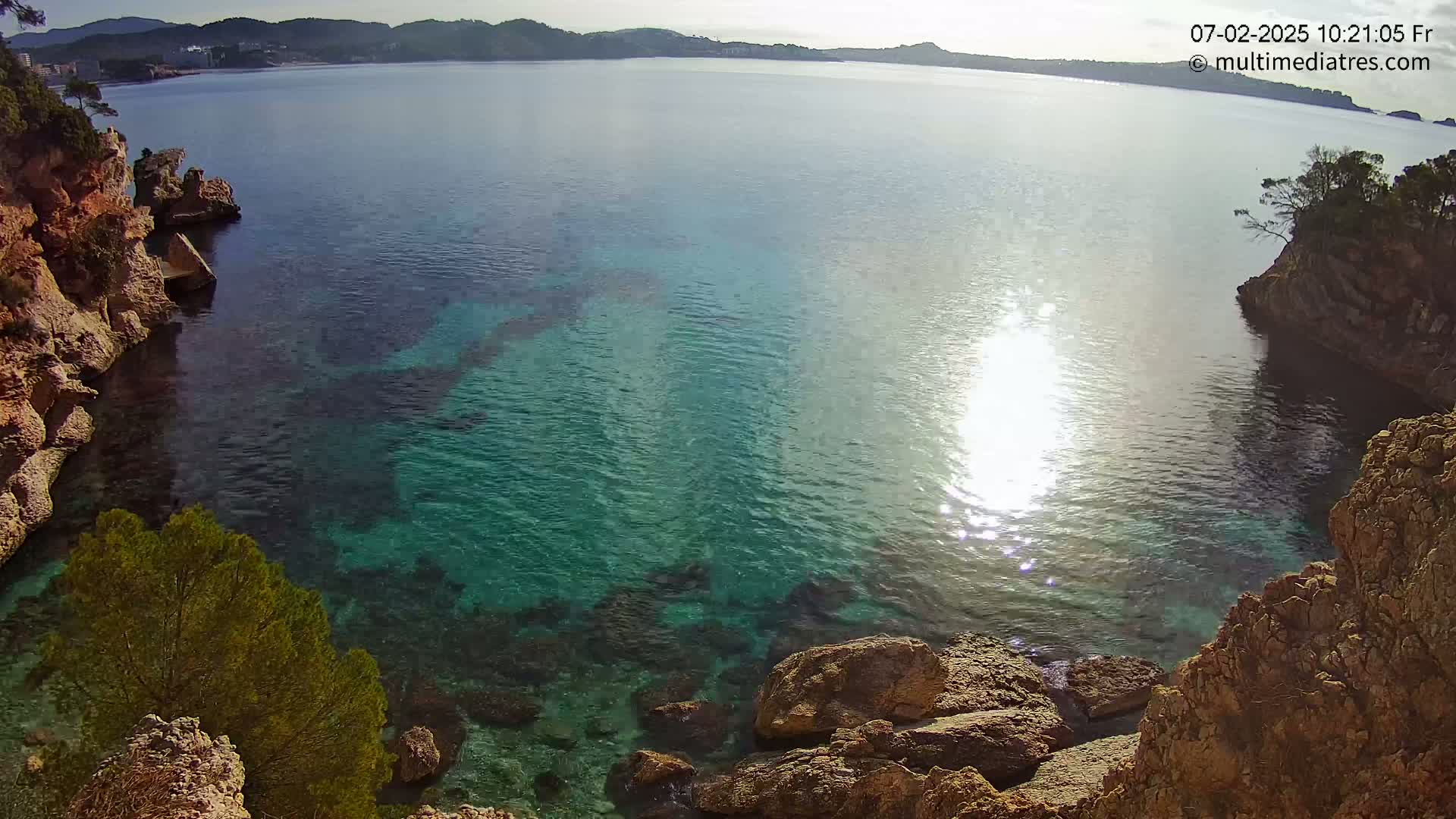 Clear, bright sunlight reflects on the calm, teal water of a secluded cove surrounded by rocky cliffs and sparse vegetation.
