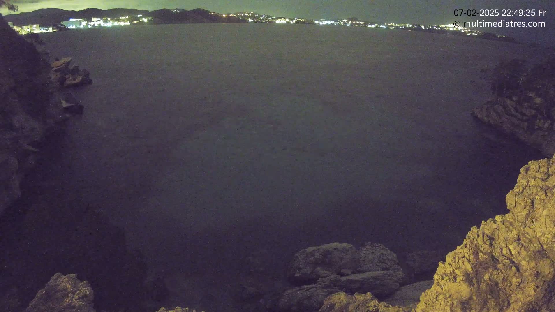 A dark, calm body of water is seen at night, nestled between rocky shorelines, with distant city lights visible on the horizon.