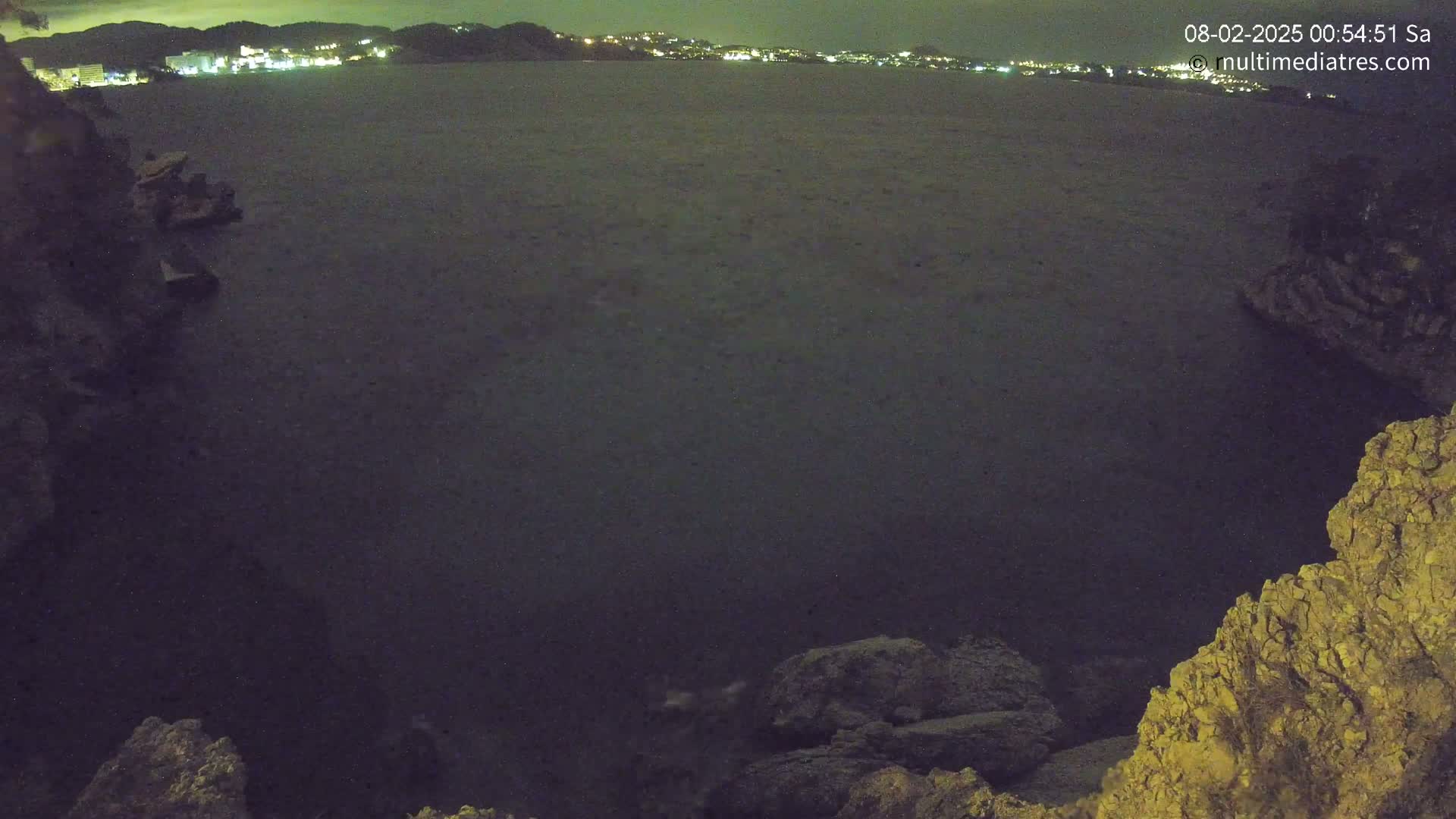A nighttime view from a rocky overlook shows a dark body of water extending to a distant, faintly lit shoreline.
