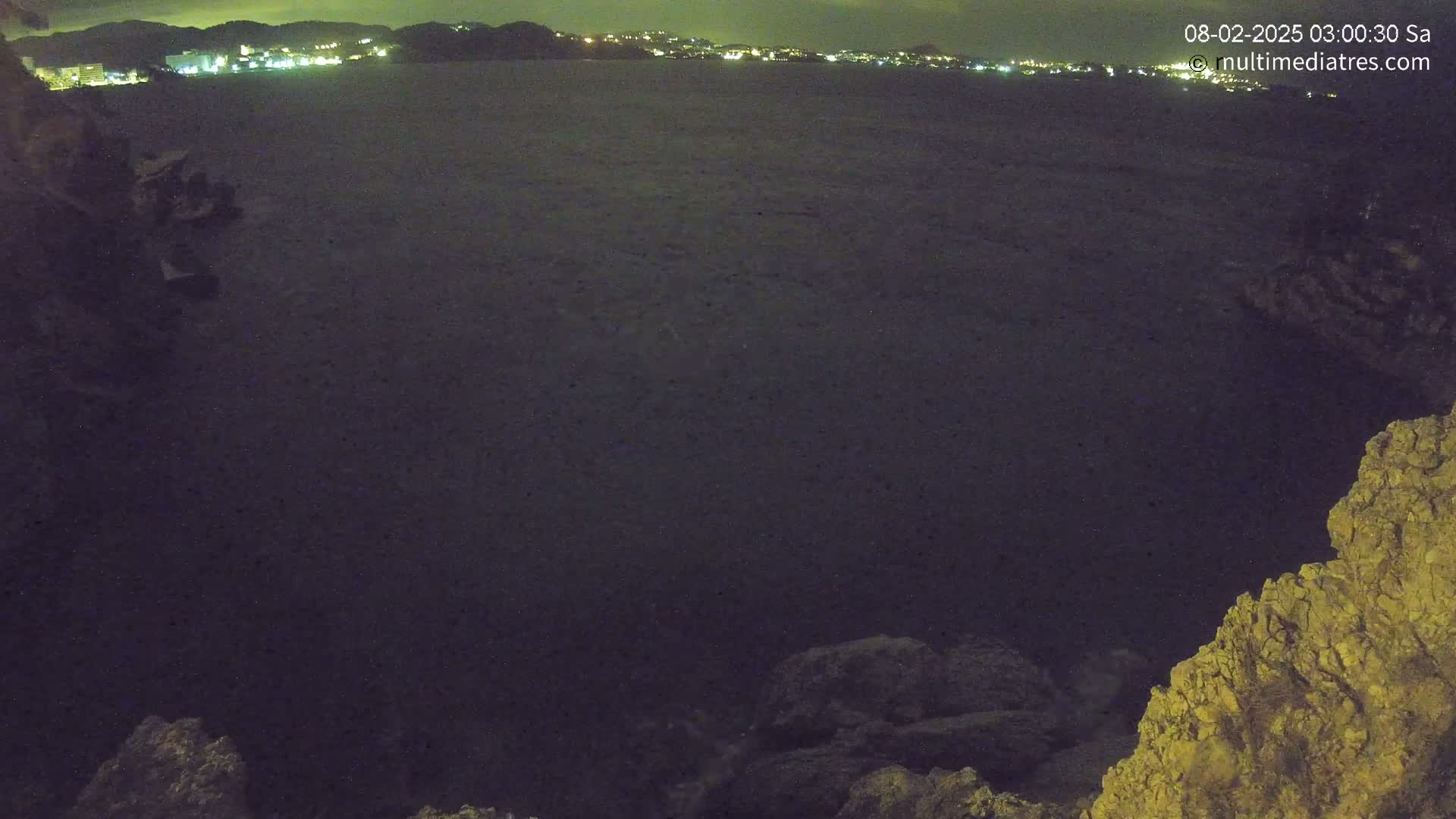 A dark, nighttime view of a calm body of water with a distant shoreline of lights and rocky foreground.