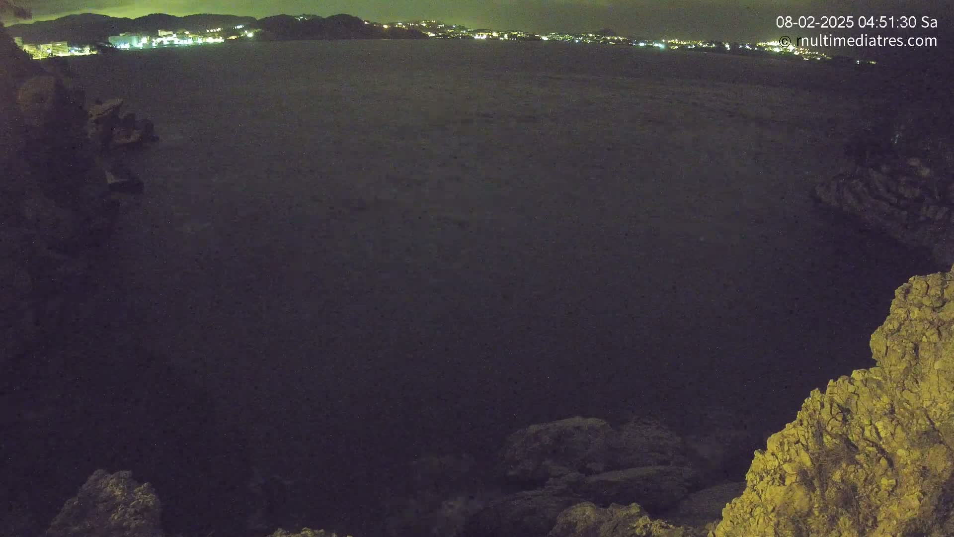 A dark, nighttime view of a calm body of water with a distant city skyline illuminated, framed by rocky terrain.