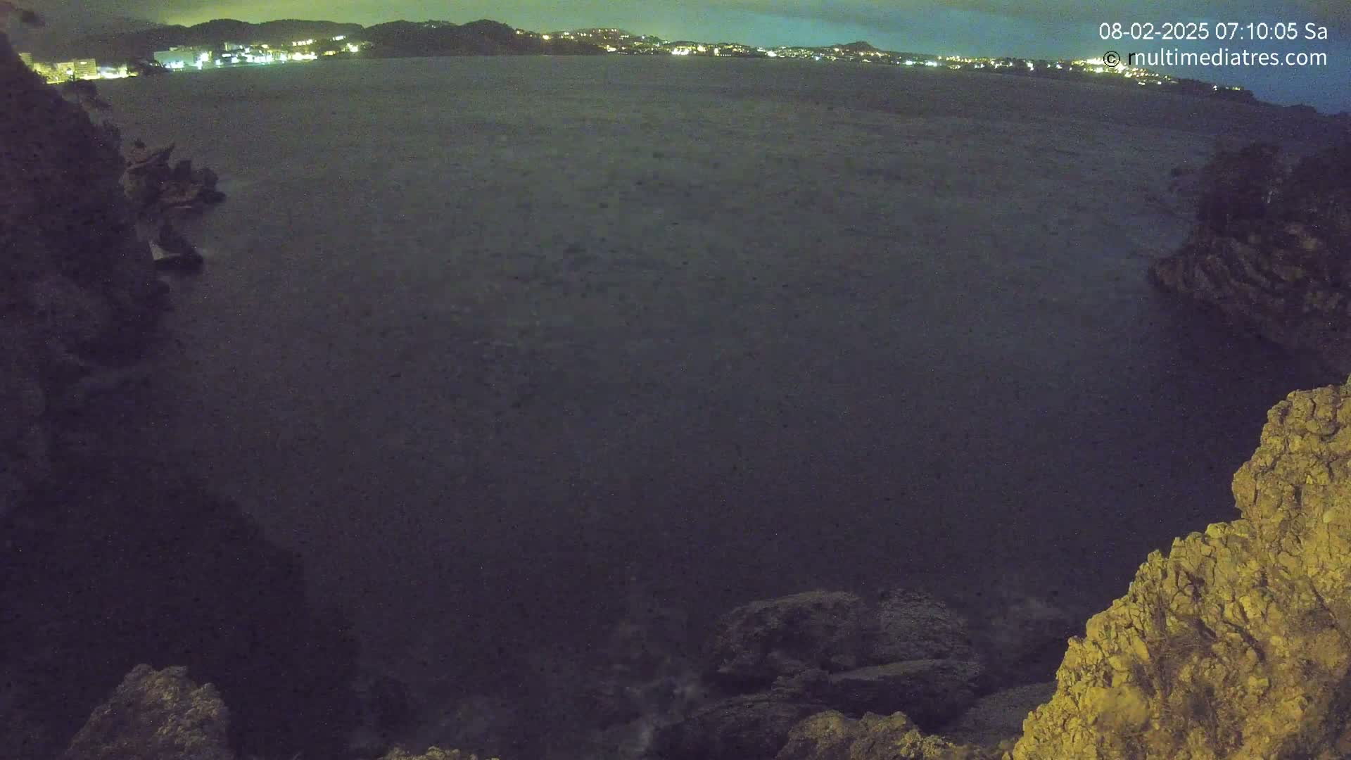 A dark, nighttime view of a calm sea from a rocky cliff overlooking a distant, illuminated shoreline.