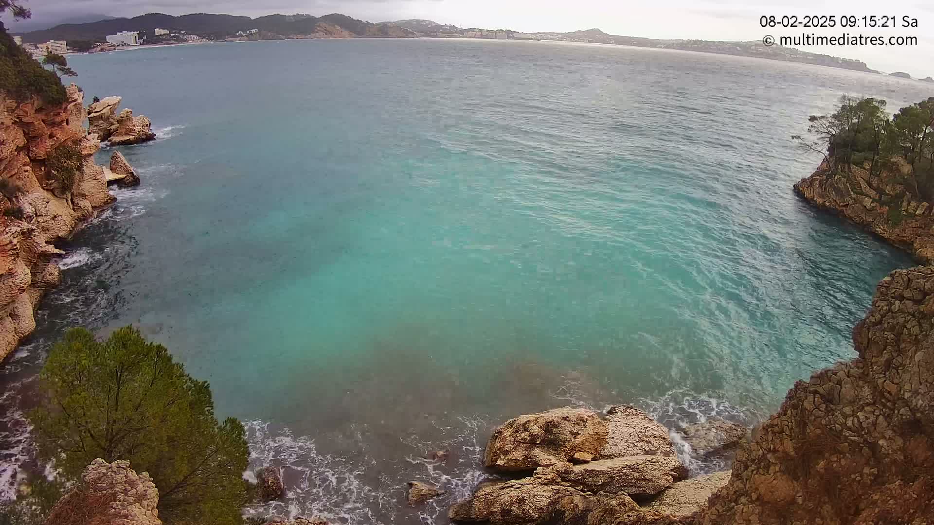 A rocky cove with turquoise water meets a calm sea under an overcast sky, overlooking a distant coastline.