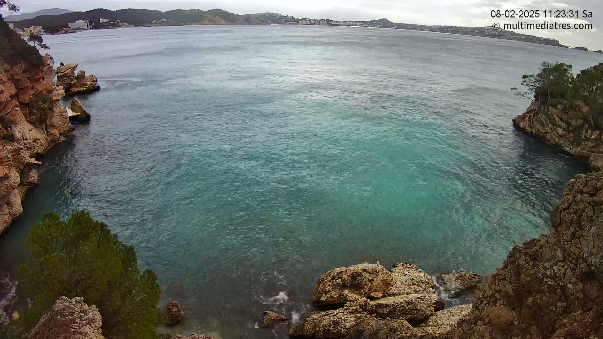 A cove of teal water is nestled between rocky, tan cliffs, with a distant coastline visible under an overcast sky.