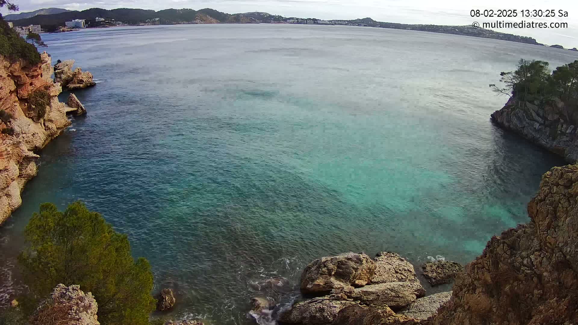A cove of clear, teal water is nestled between rocky, cliff-like shores under a mostly cloudy sky.
