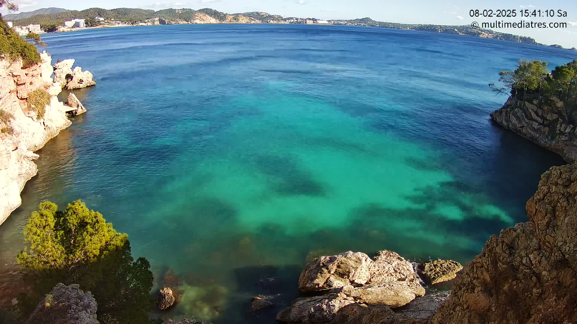 Clear, turquoise water fills a cove surrounded by rocky, light-brown cliffs and greenery under a sunny sky.