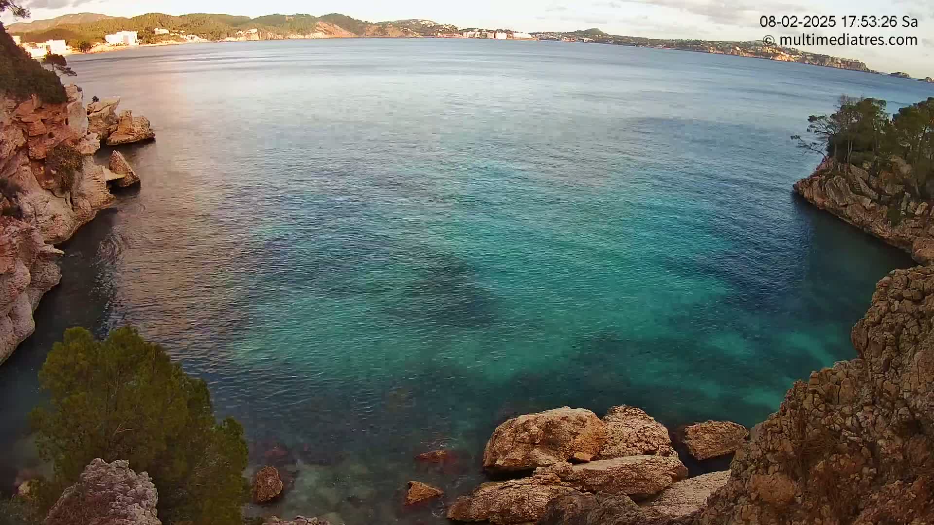 A tranquil cove of clear, teal water is nestled between rocky, tan cliffs, with a distant shoreline visible under a partly cloudy sky.