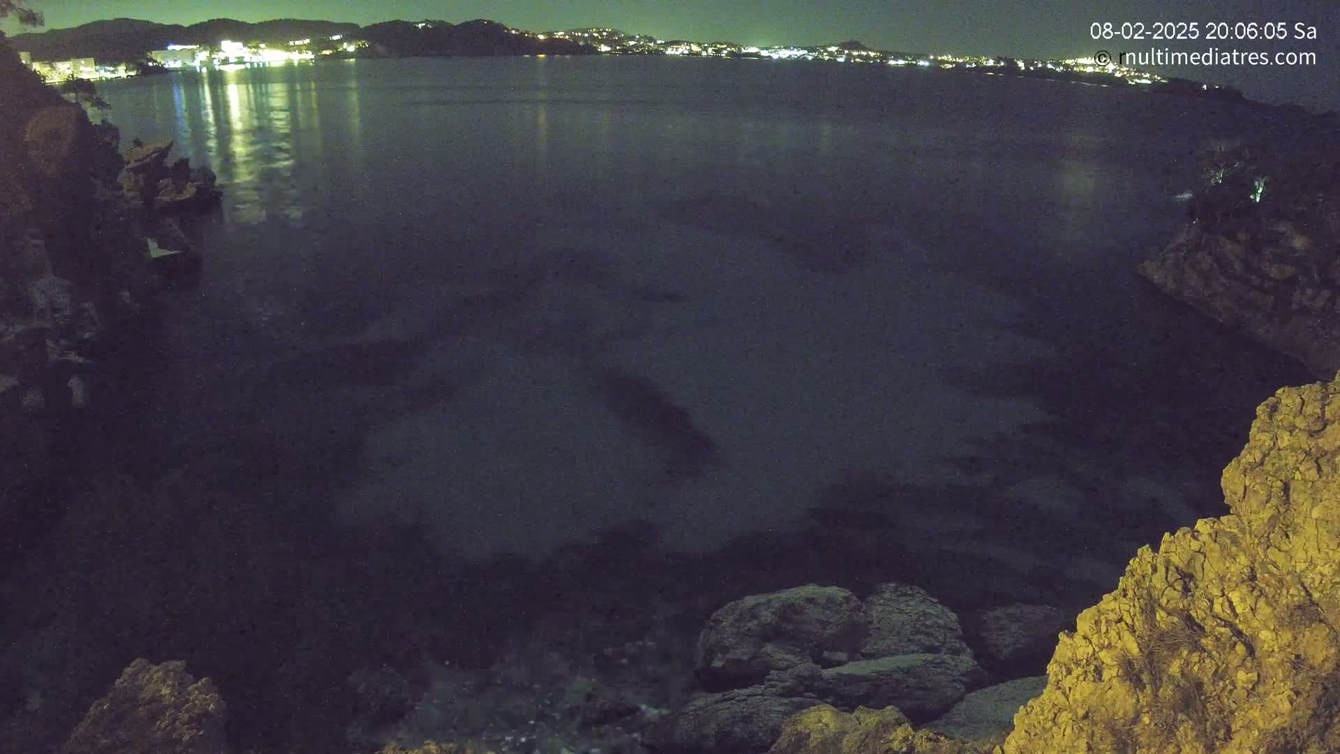 A nighttime view of a dark body of water reflecting the lights of a city on the opposite shore, from a rocky vantage point.