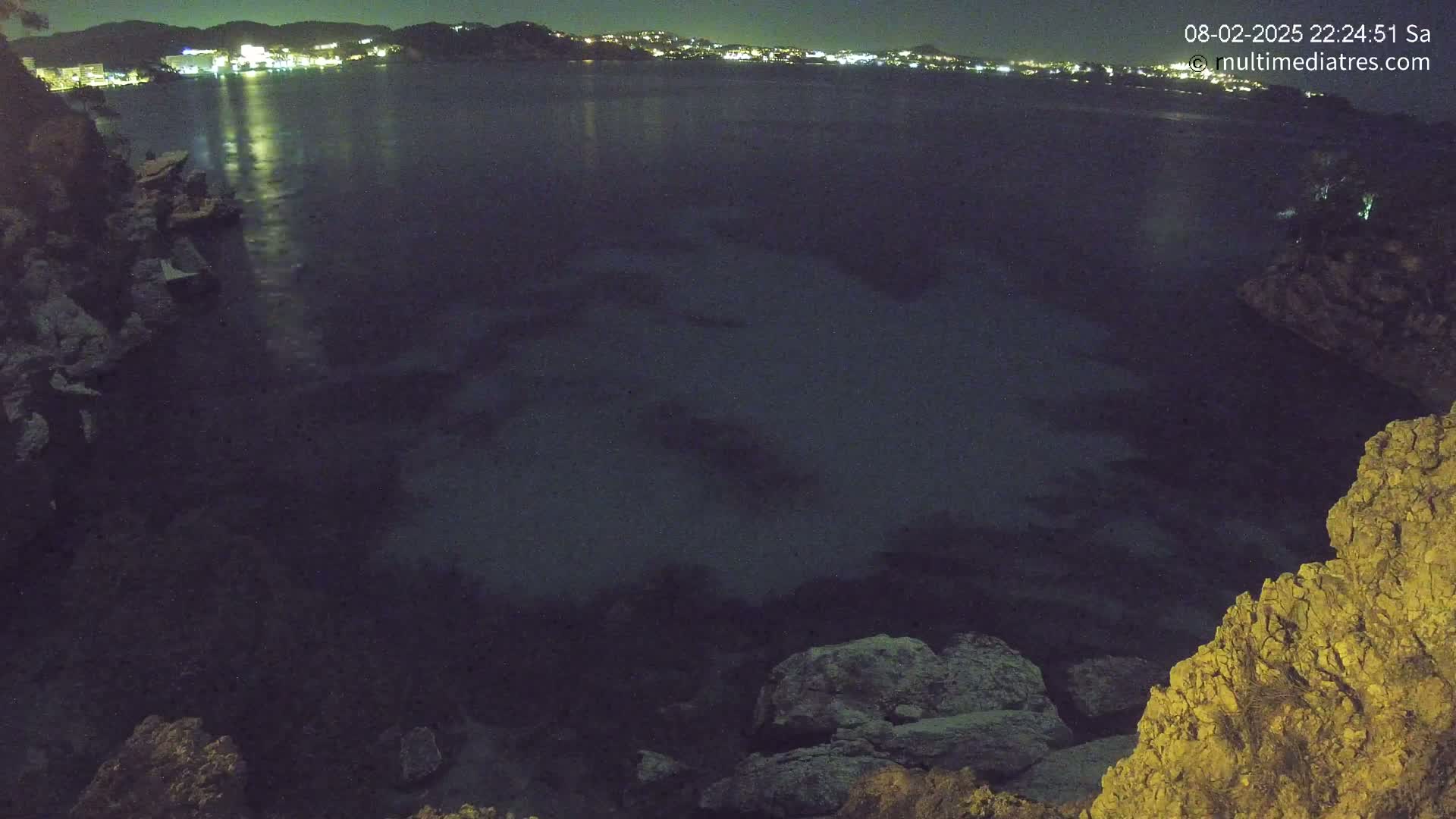 A nighttime view of a dark body of water with rocky shorelines, and a distant city illuminated with lights.
