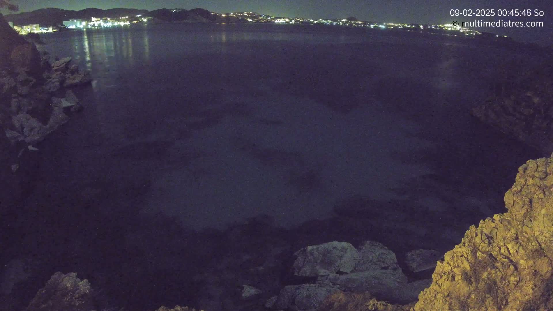 A nighttime view of a dark body of water, reflecting the lights of a distant town, seen from a rocky outcrop.