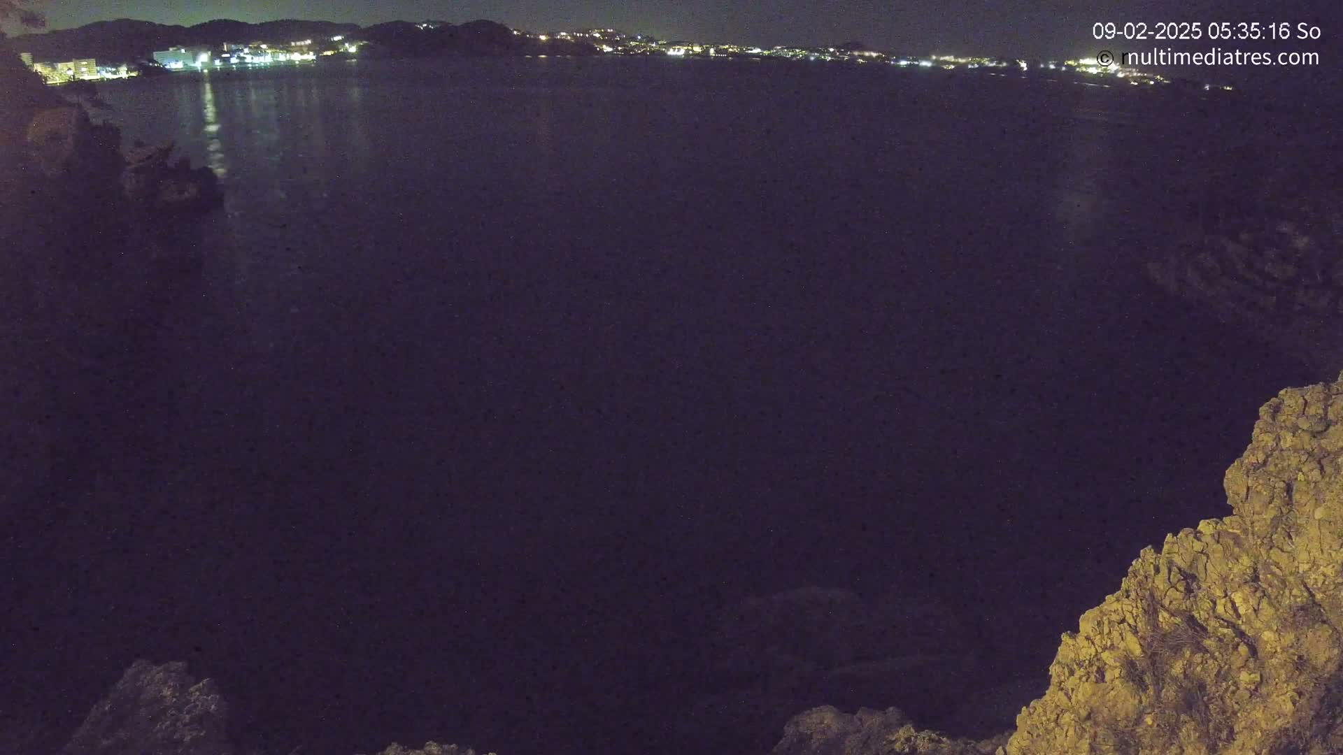 A nighttime view of a dark body of water reflecting the lights of a distant town, seen from a rocky cliff.