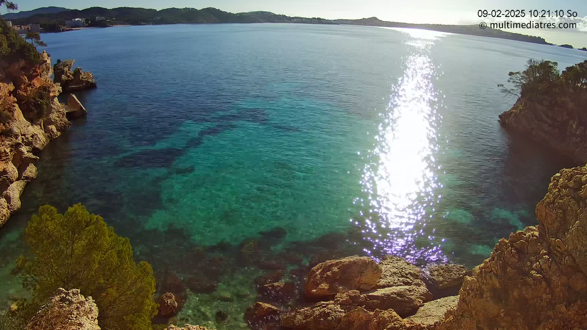 Clear, sunny day overlooking a calm, bright turquoise ocean cove surrounded by rocky cliffs and sparse vegetation.