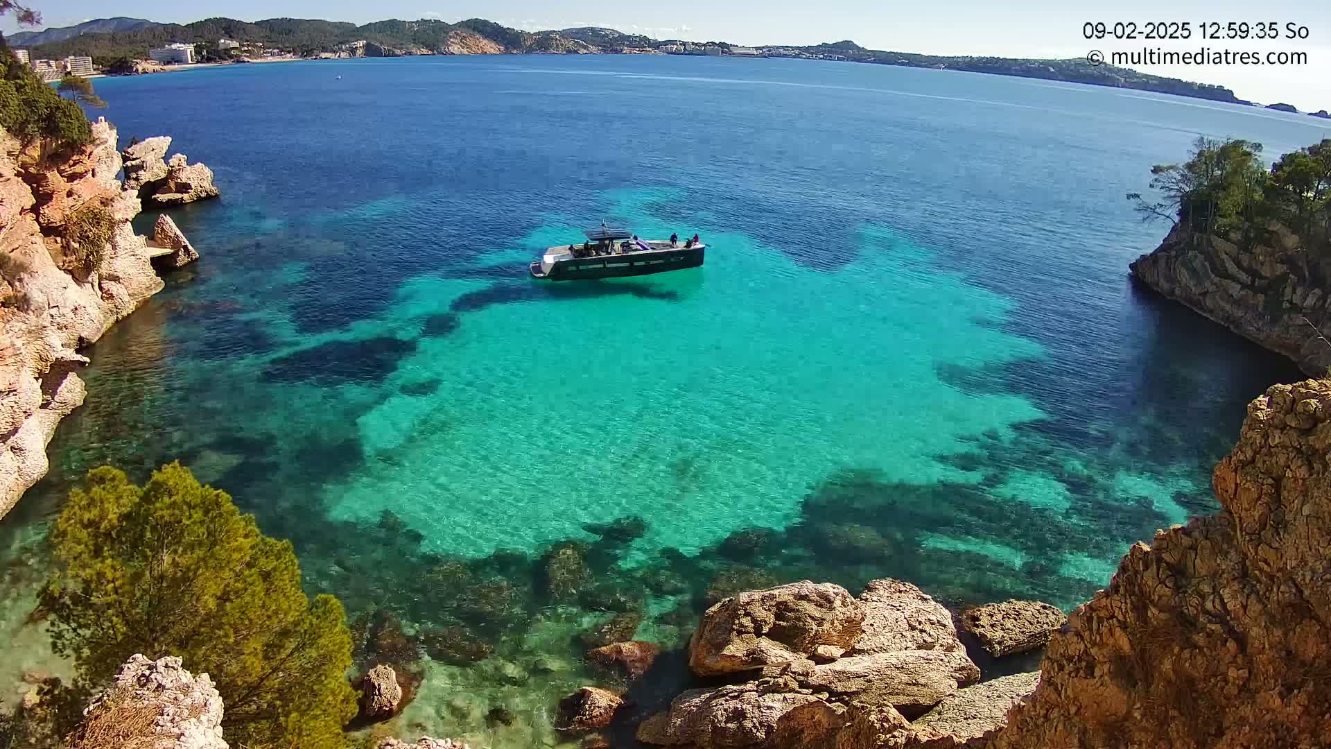 A dark-colored motorboat is anchored in a calm, clear turquoise cove surrounded by rocky, tan-colored cliffs on a sunny day.