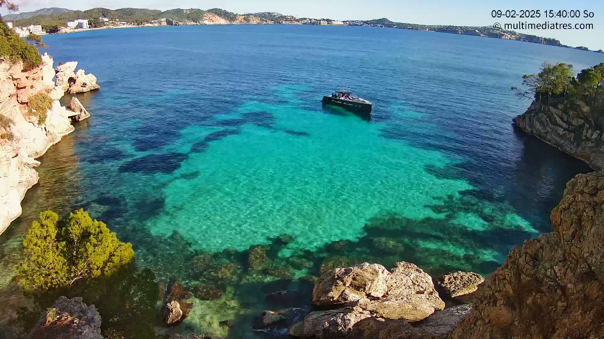 A small boat is in a cove of clear, turquoise water surrounded by rocky, light-brown cliffs on a sunny day.