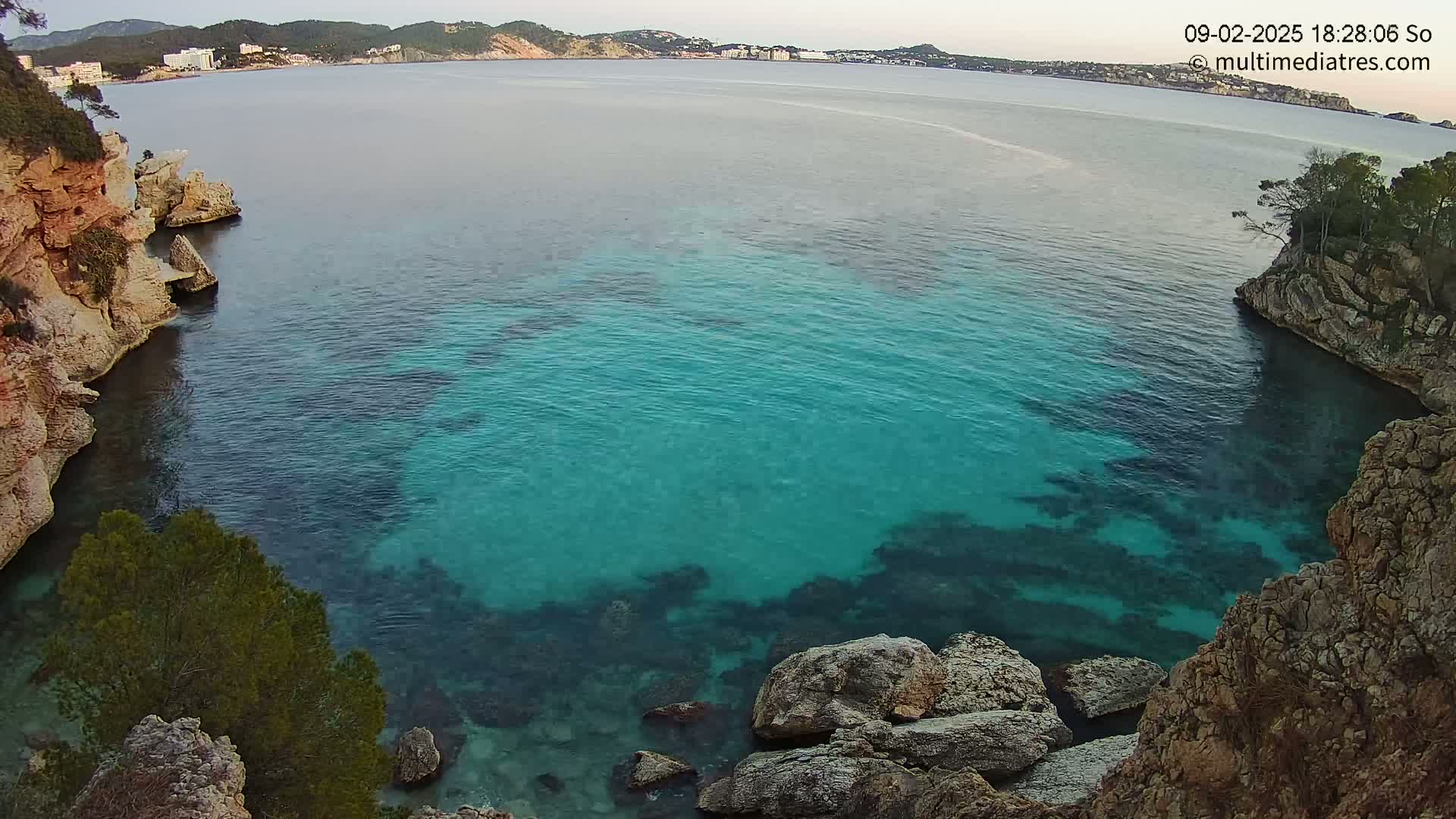A cove of clear, teal water is nestled between rocky cliffs, with a distant view of a coastline under a calm sky.