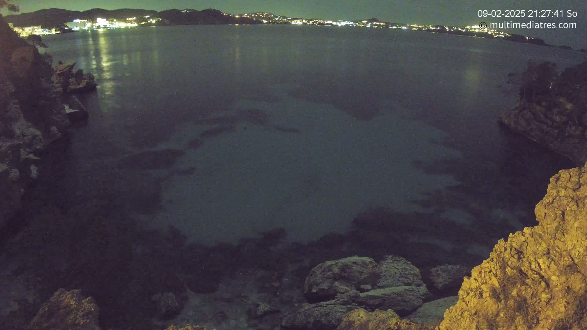A nighttime view of a dark body of water, illuminated by distant city lights, from a rocky vantage point.