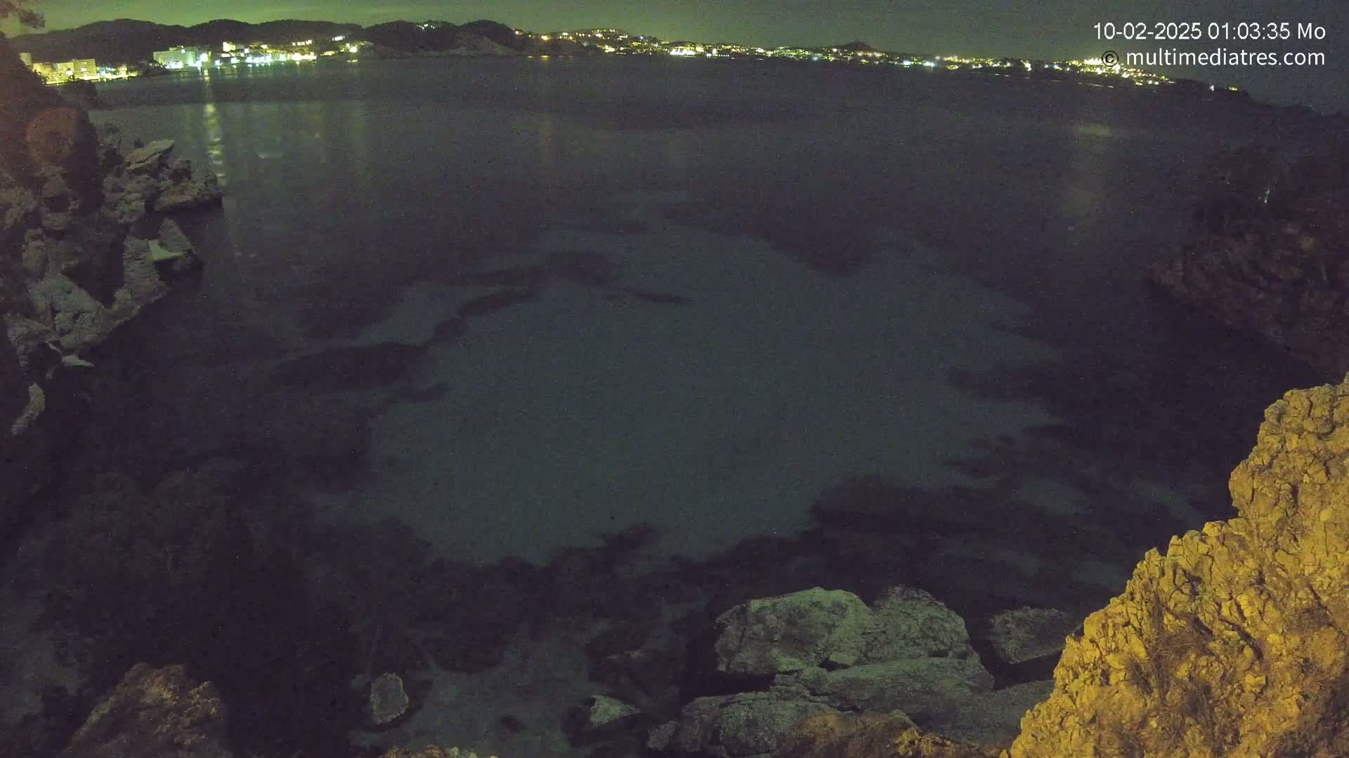 A nighttime view of a dark, calm body of water surrounded by rocky shores, with a distant town illuminated by lights.
