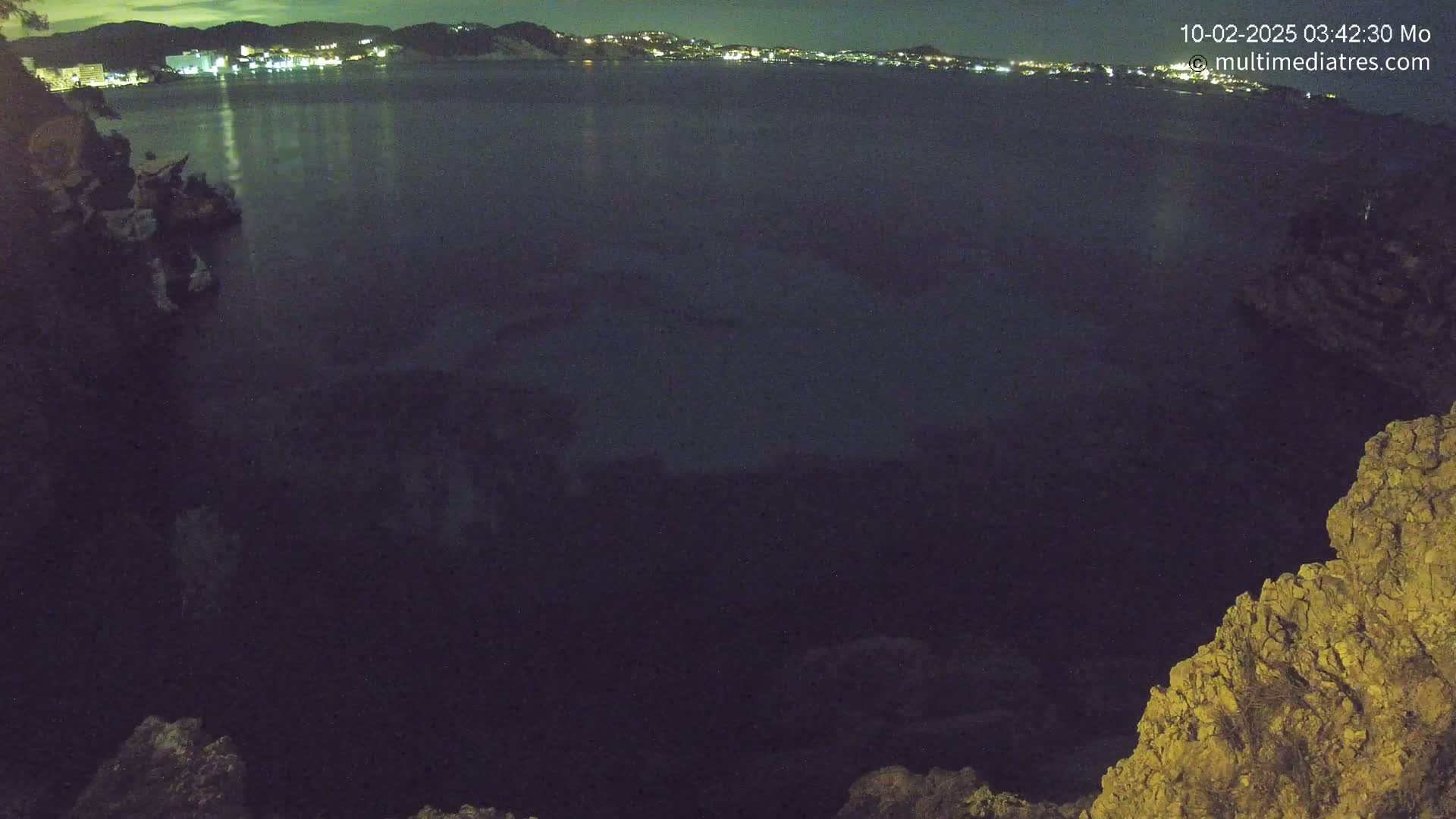 A nighttime view of dark water, possibly a cove or bay, with a distant, illuminated shoreline and rocky cliffs in the foreground under a clear sky.