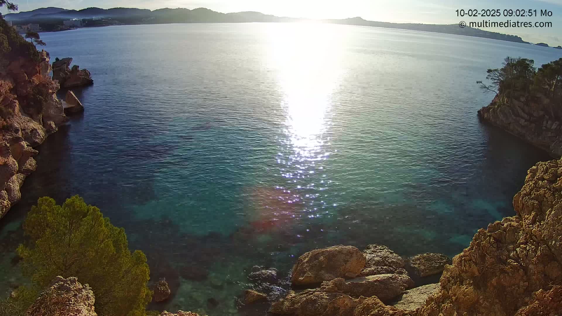 Rocky cliffs surround a calm, clear cove of turquoise water reflecting the bright sunlight of a sunny morning.