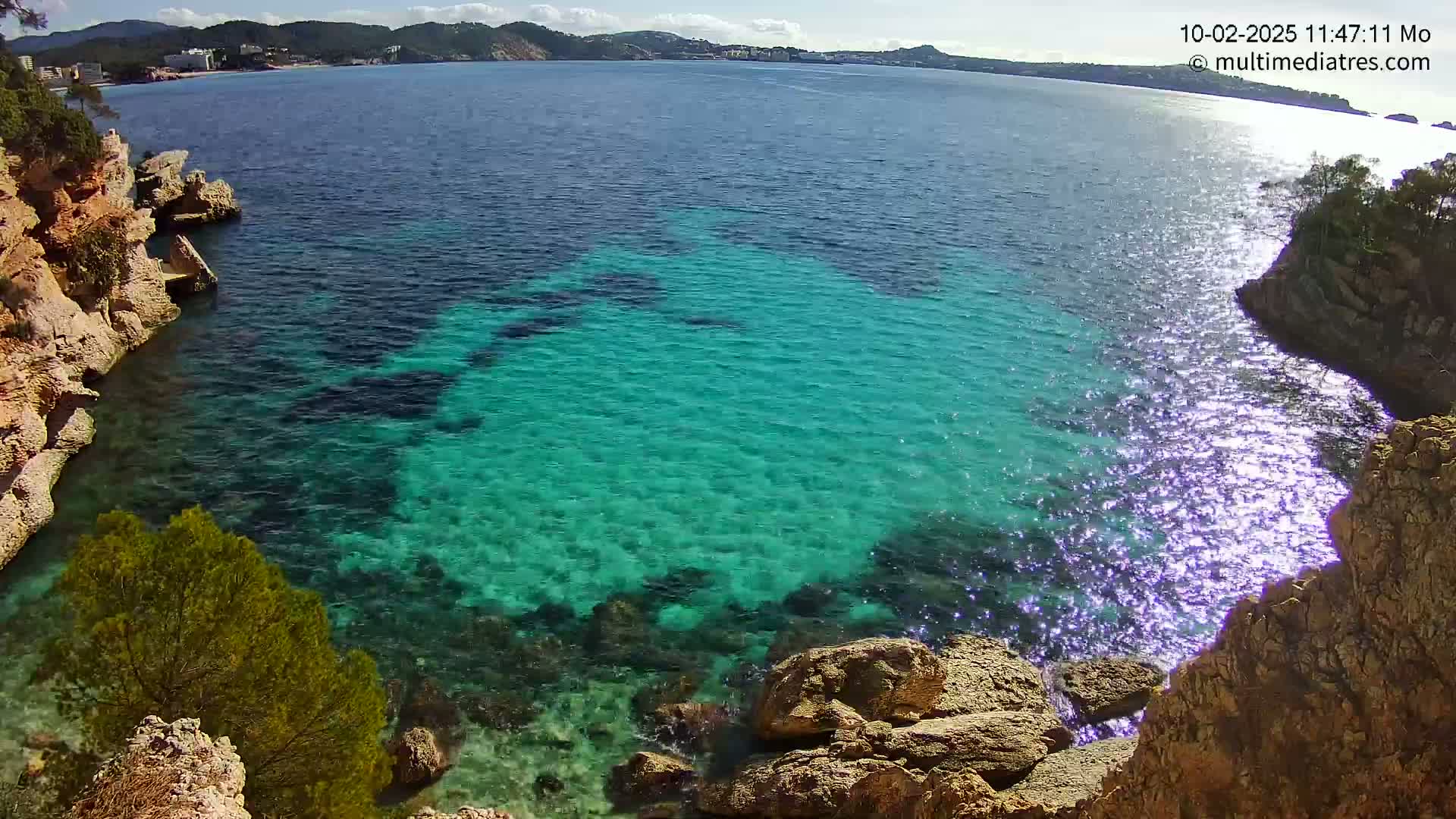 A rocky cove with vibrant turquoise water under a sunny sky shows a distant shoreline.