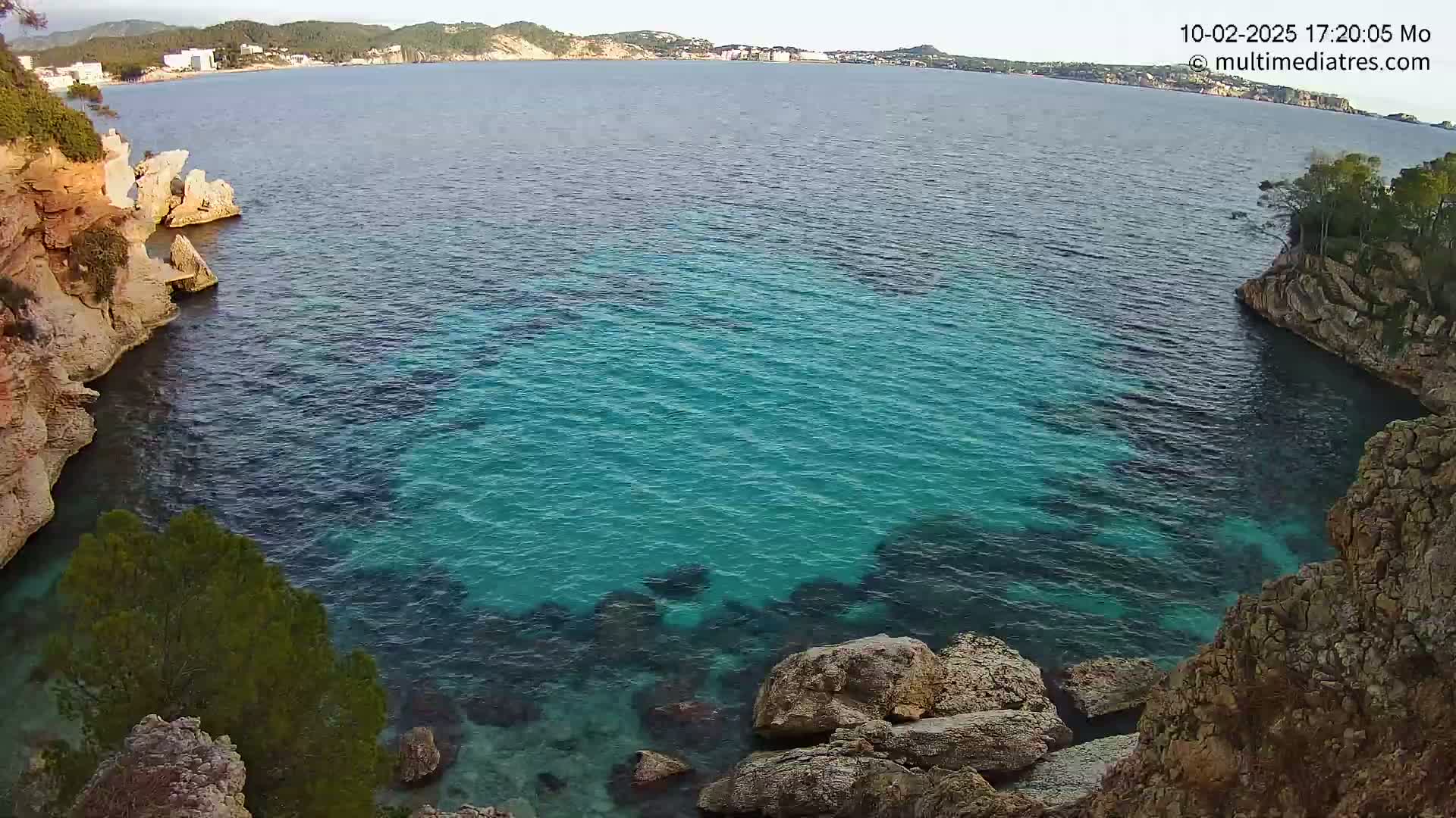 A secluded cove with clear, turquoise water is nestled between rocky cliffs, overlooking a calm sea and distant coastline under a clear sky.