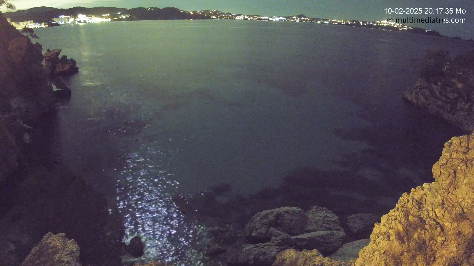 A dark, calm body of water is seen at night, reflecting lights from a distant town on the horizon, with rocky cliffs visible in the foreground.