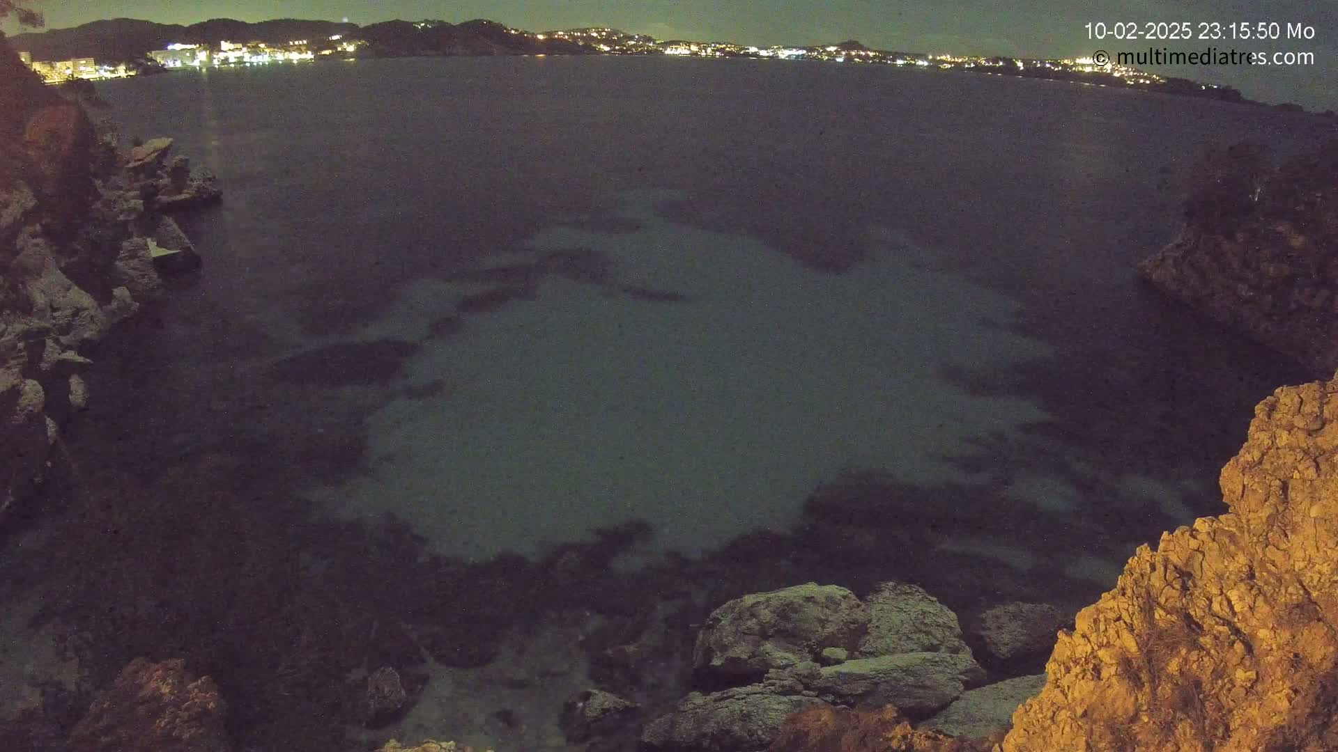 A nighttime view of dark, calm water, partially illuminated, near a rocky shoreline with a city faintly visible in the distance.