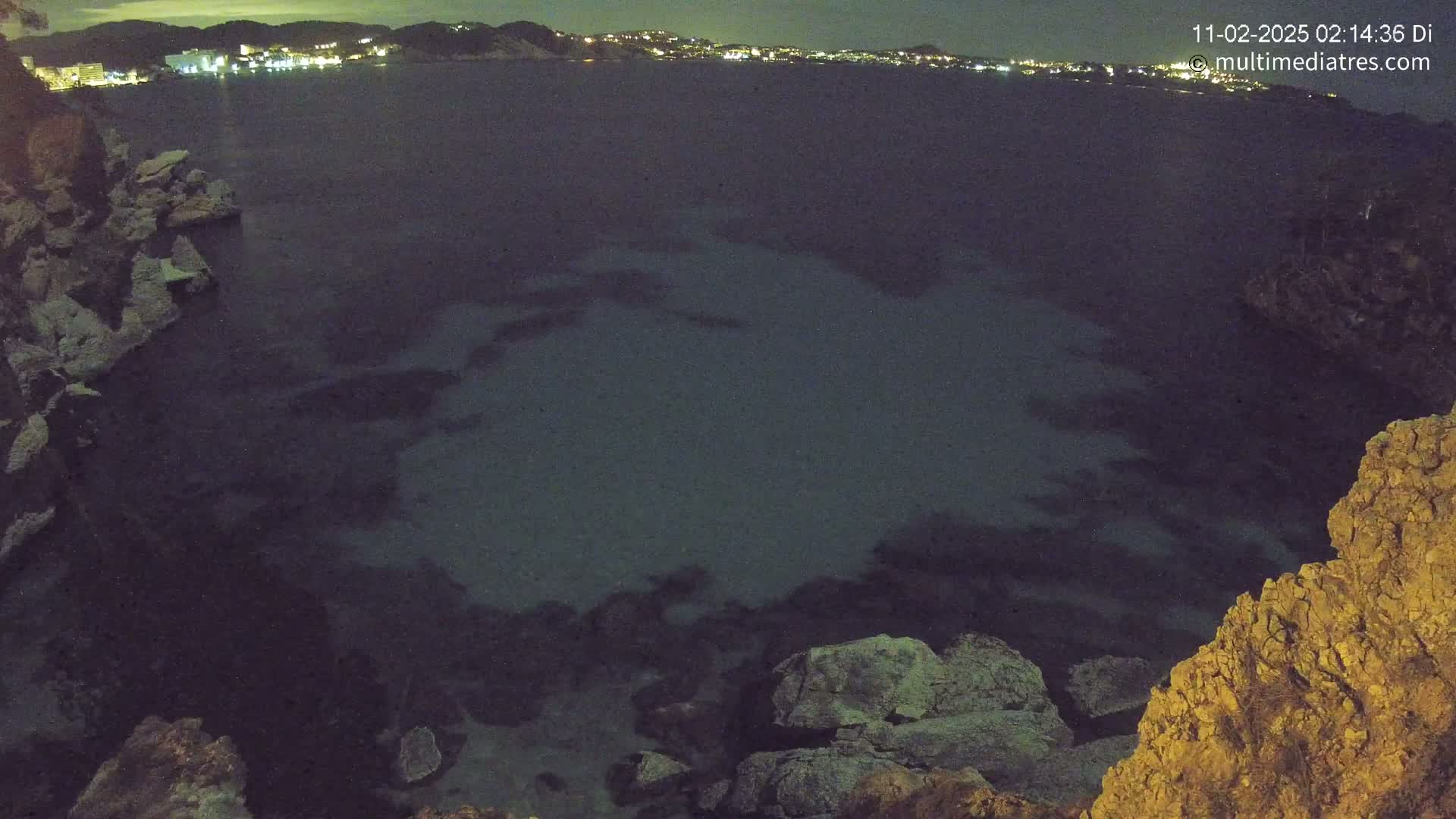 A dark, nighttime view of a calm body of water next to rocky shorelines, with a distant town illuminated by lights.