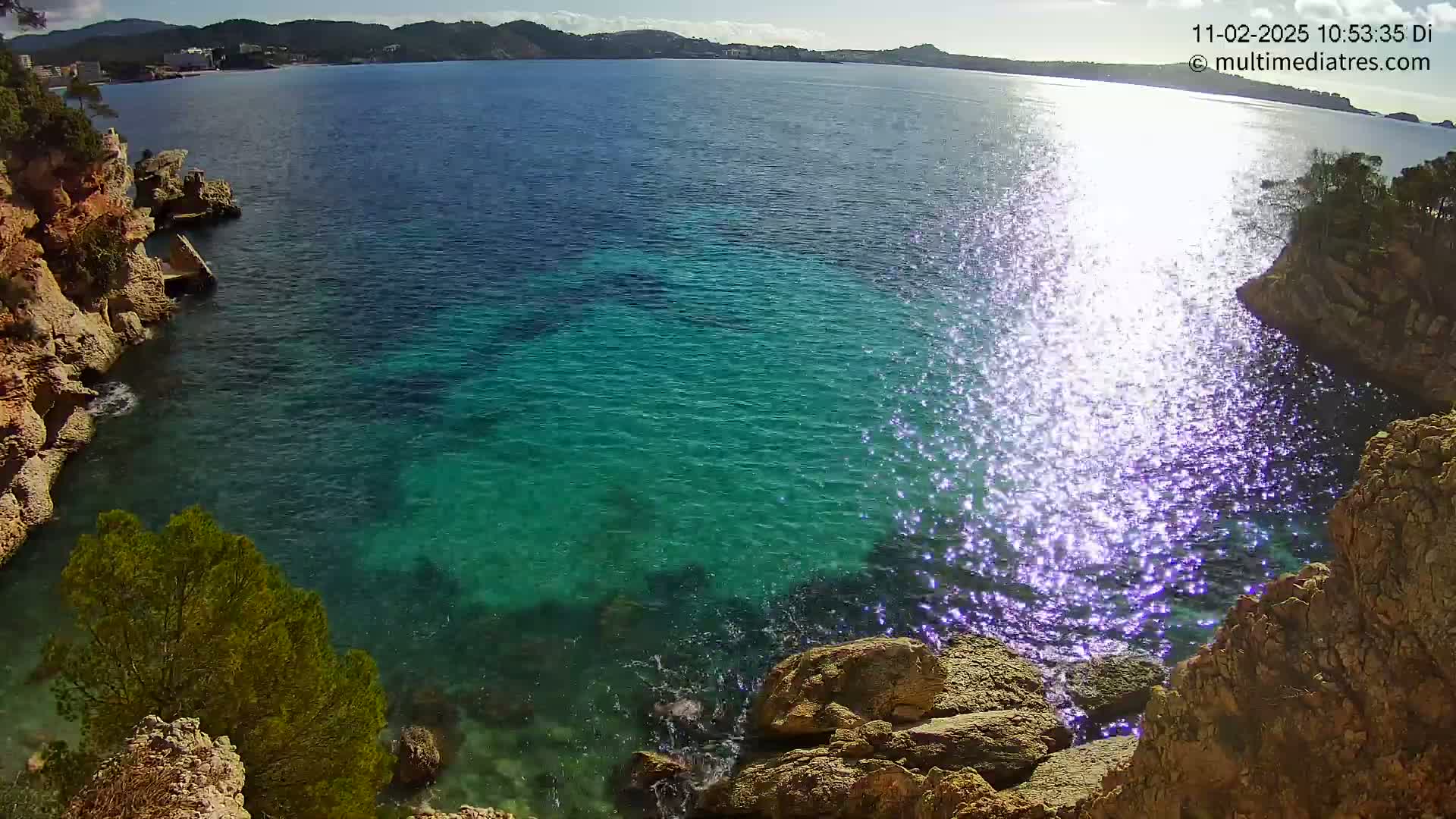 Clear, bright sunlight glistens on the surface of a calm, teal ocean nestled between rocky shores.