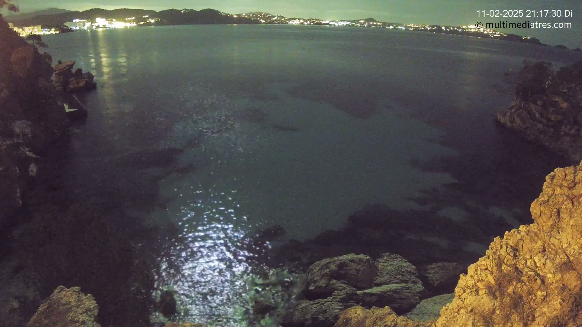 A nighttime view of a dark, calm body of water reflecting city lights across from a rocky shoreline.