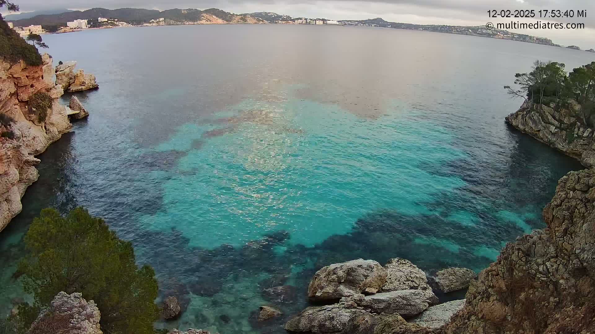 A cove of clear, teal water is nestled between rocky, beige cliffs on an overcast day, with a distant shoreline visible across the water.