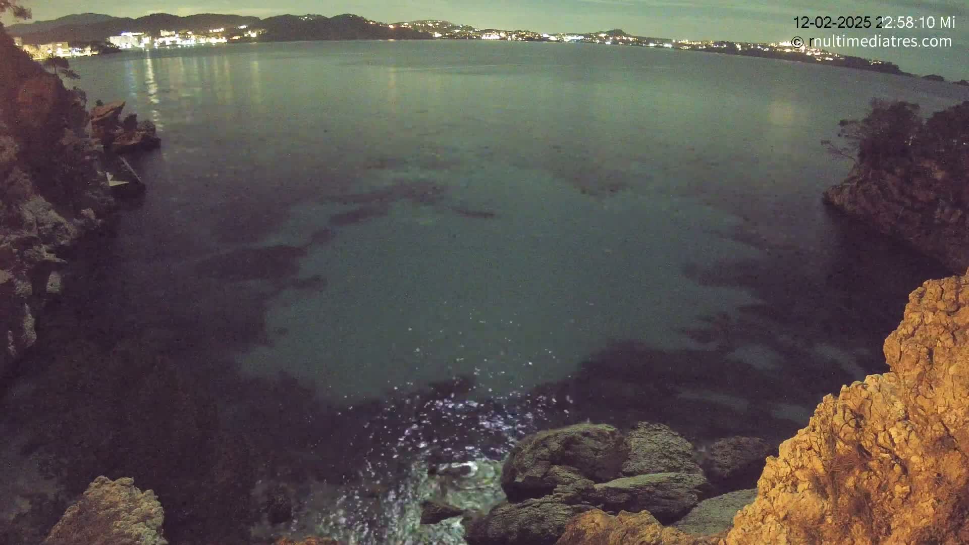 A nighttime, high-angle view shows dark, calm water between rocky coastlines, with a town faintly lit in the distance.